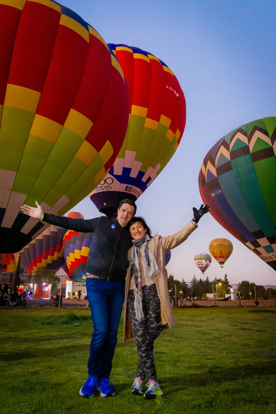 Couple with arms outstretched before vivid hot air balloons at blue hour over Teotihuacan