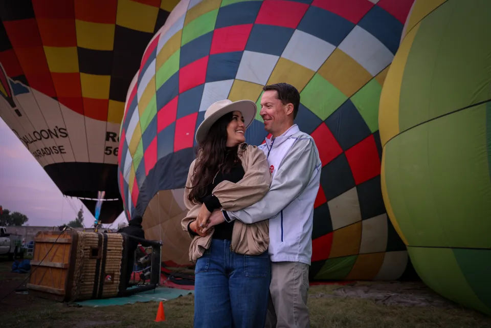 Couple gazing at each other beside colorful hot air balloons inflating at dawn