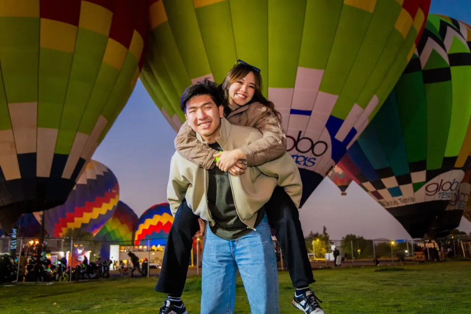 Playful couple piggyback riding in front of glowing hot air balloons at dawn