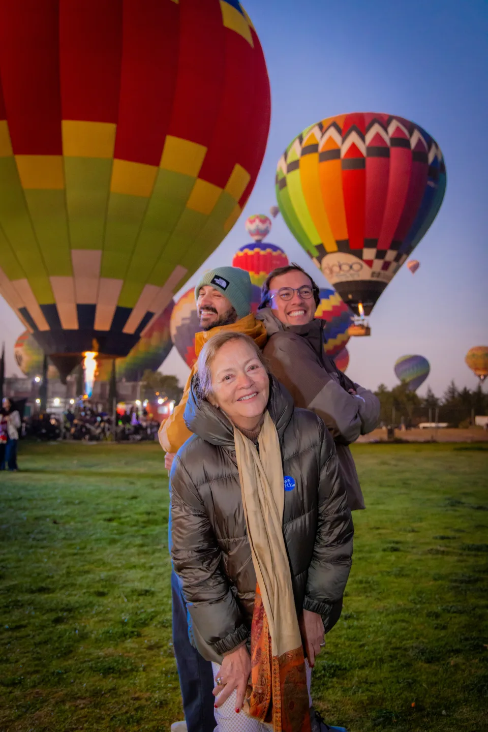 Mother carrying her two grown sons on her back laughing before glowing hot air balloons at dawn