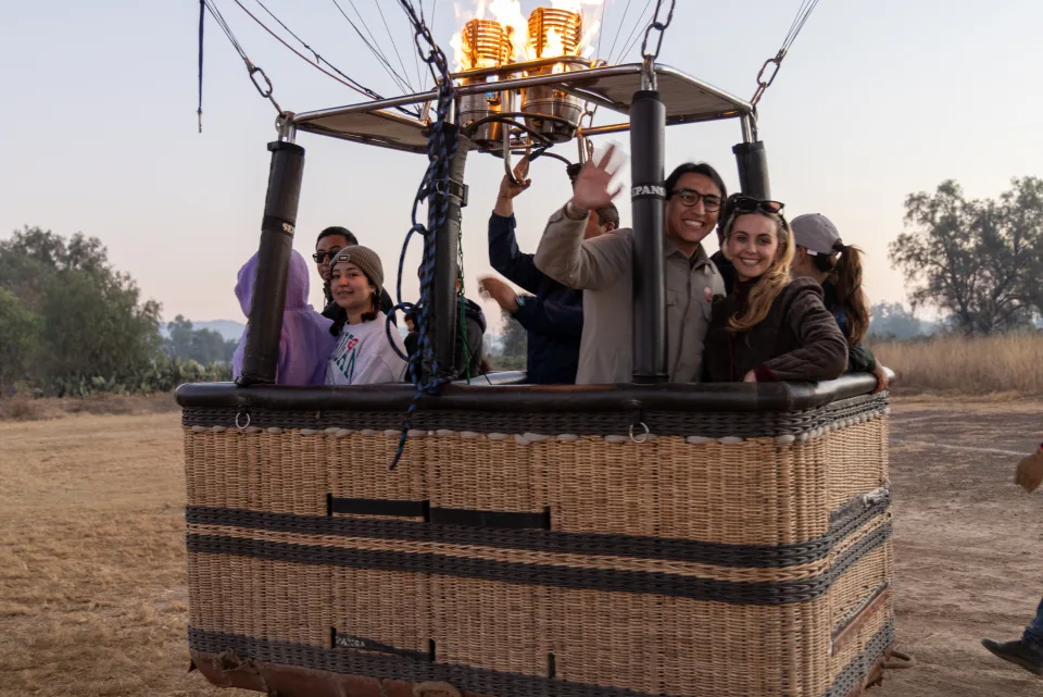 Group of passengers waving from a hot air balloon basket before sunrise liftoff