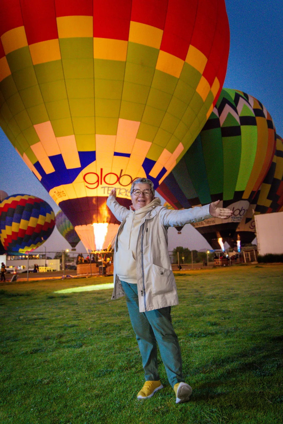 Joyful older woman with arms wide open before glowing hot air balloons at dawn