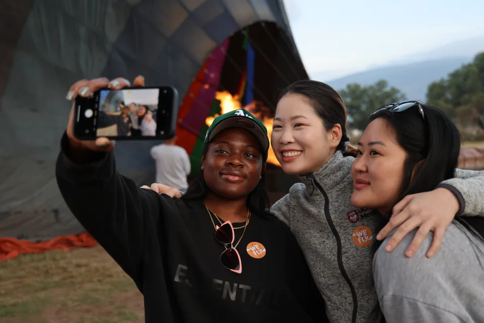 Three friends taking a selfie beside a glowing hot air balloon at dawn