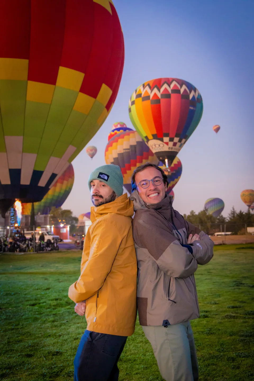 Two friends posing back-to-back with colorful hot air balloons at sunrise