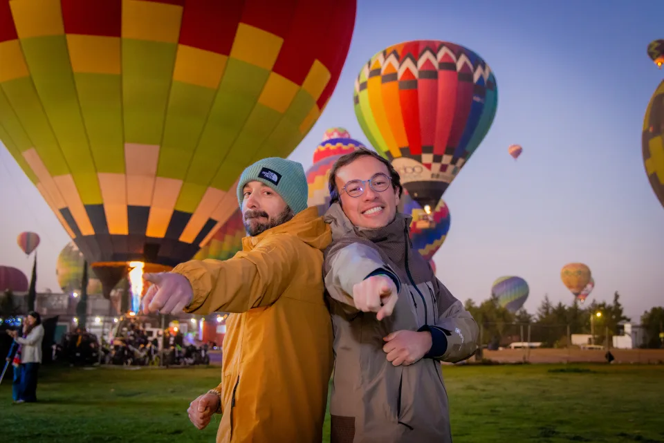 Two friends pointing at the camera with sunrise hot air balloons behind them