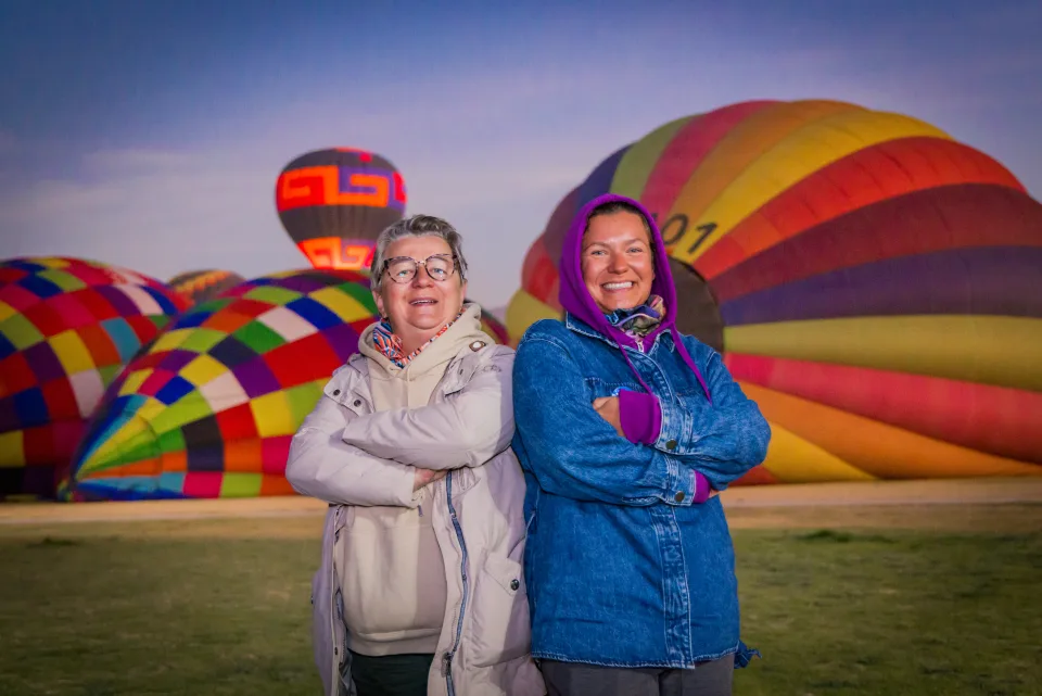 Two women posing back-to-back with colorful balloons inflating at dawn