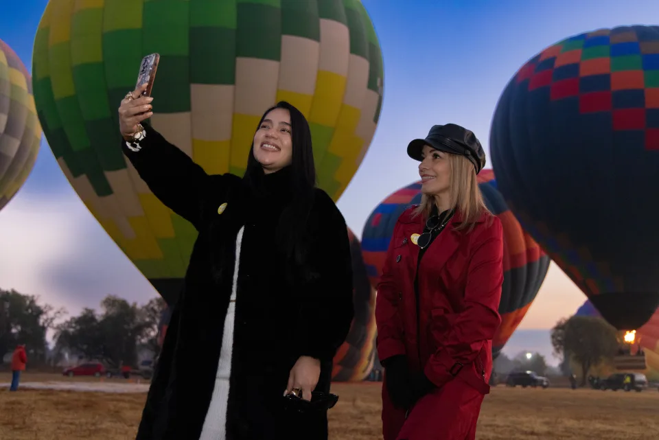 Two women taking a selfie with hot air balloons in the field at first light
