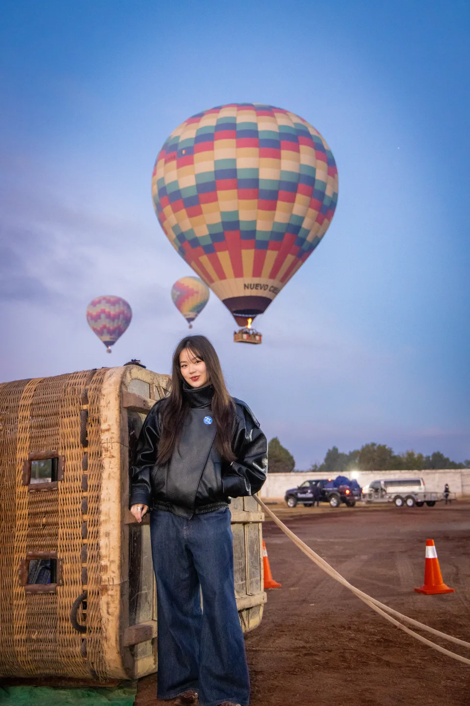Woman standing beside a balloon basket as another balloon floats overhead at dawn