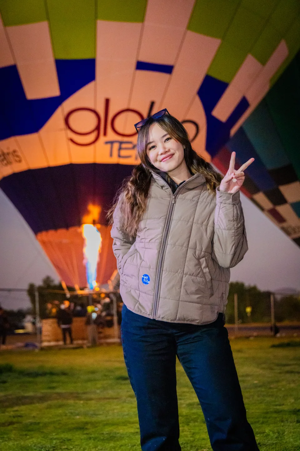 Young woman making a peace sign before a glowing hot air balloon and burner flame