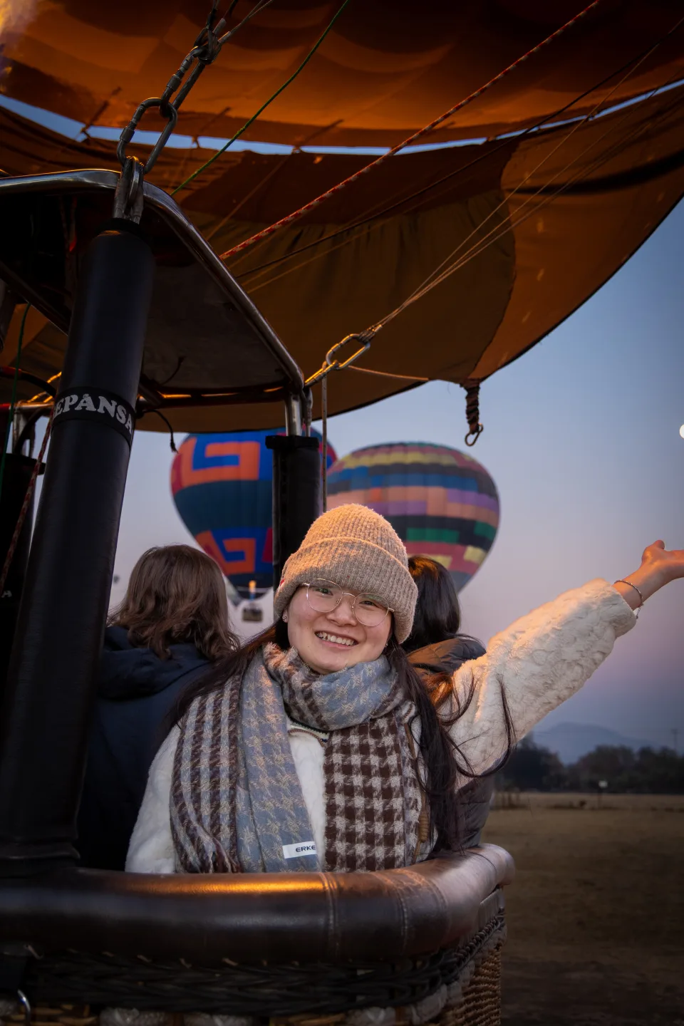 Bundled-up woman smiling in the basket with balloons drifting behind at dawn