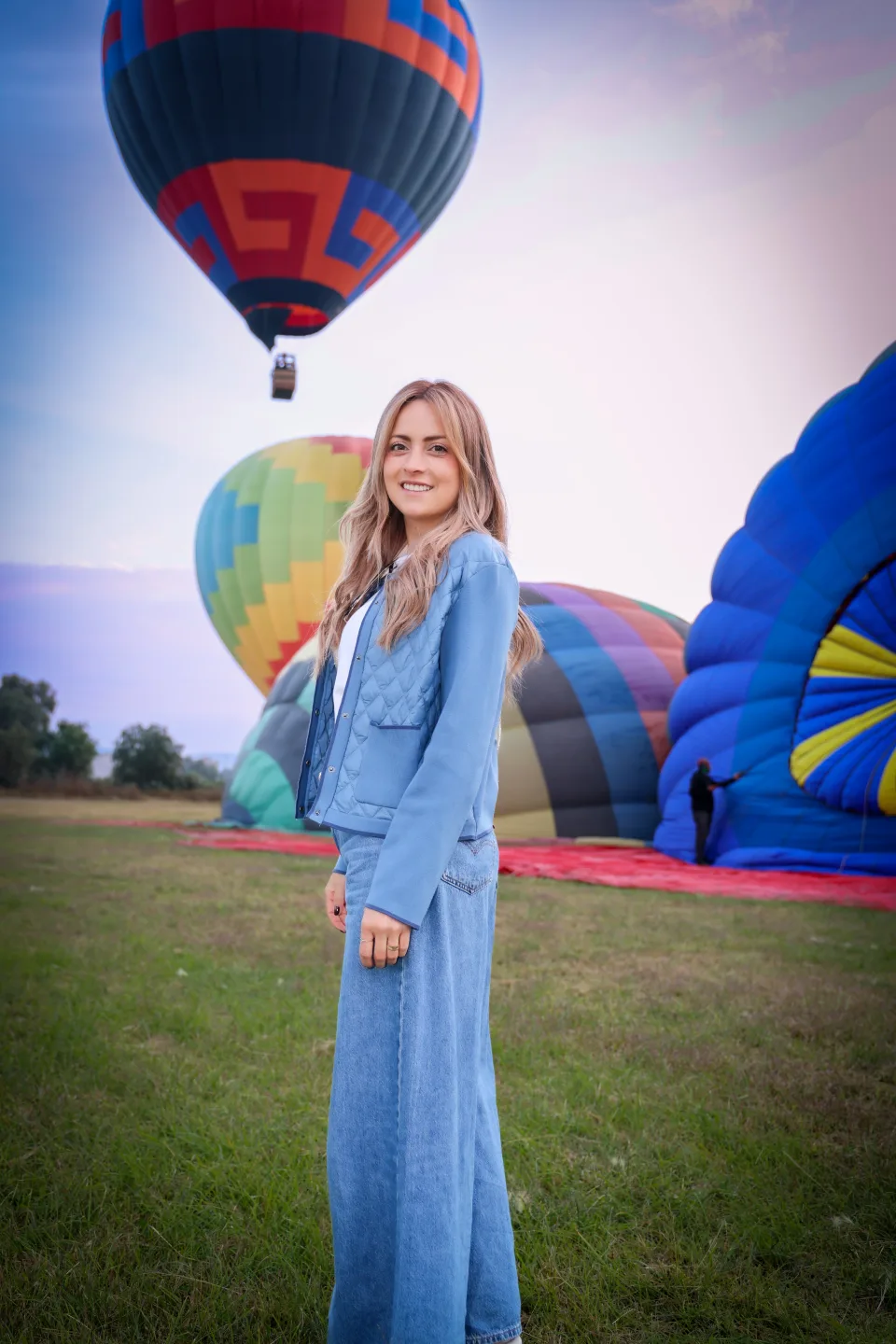 Woman standing in the launch field with a hot air balloon rising behind her at dawn